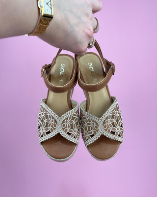 Pair of brown sandals with white lace details held by a hand against a pink background