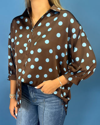 A woman wearing a brown button-up blouse with large light blue polka dots, paired with blue jeans, standing against a solid blue background.