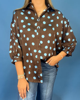 A woman wearing a brown button-up blouse with light blue polka dots, paired with blue jeans, standing against a solid blue background.