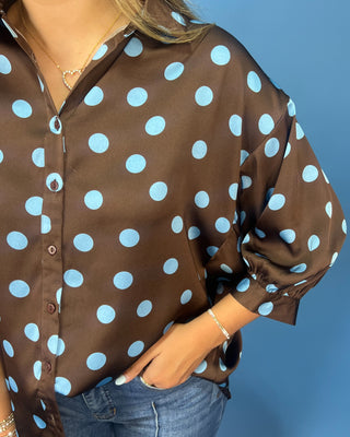 A woman wearing a brown button-up blouse with large light blue polka dots, paired with blue jeans, shown against a solid blue background.