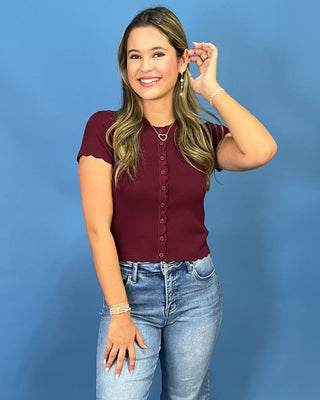 A woman modeling a short-sleeve burgundy button-up top with scalloped sleeves, paired with light-wash blue jeans, standing against a solid blue studio background.