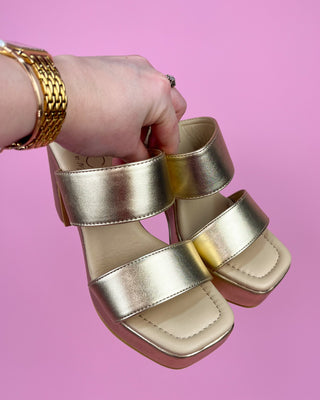 Gold sandals held by a hand against a pink background