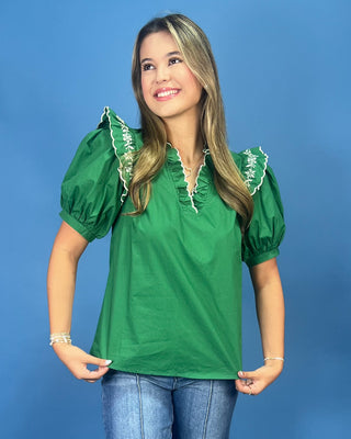 A woman modeling a green short-sleeve blouse with puff sleeves, white ruffle trim, and a white embroidered V-neckline, paired with blue jeans against a solid blue background.