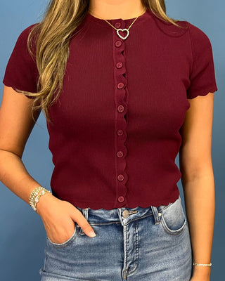A woman wearing a short-sleeve ribbed knit top in deep burgundy with a scalloped button-front closure, paired with blue jeans against a solid blue background.
