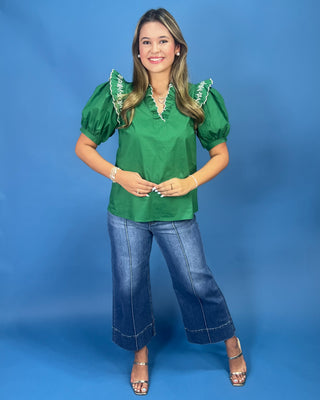 A woman modeling a green short-sleeve blouse with ruffled trim at the neckline and cuffs, paired with wide-leg blue jeans and metallic heeled sandals, against a solid blue studio background.