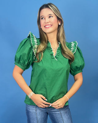 A woman wearing a bright green short-sleeve blouse with white embroidered ruffle trim at the collar and shoulders, paired with blue jeans, standing against a solid blue studio background.