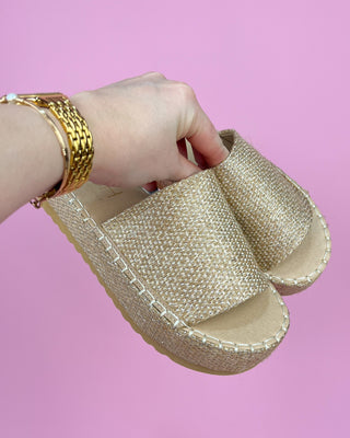 Gold glittery sandal held by a hand against a pink background