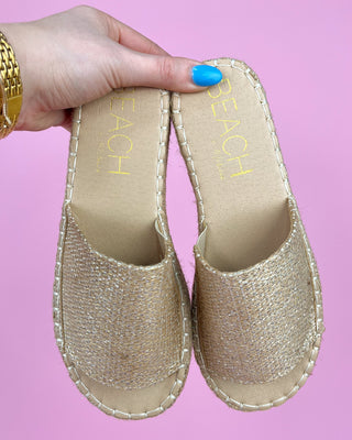 Gold espadrille sandals with 'Beach' text on a pink background