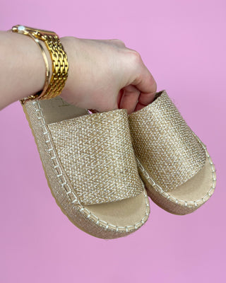 Gold espadrille sandals held by a hand against a pink background