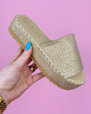 Gold espadrille sandal held by a hand with blue nail polish against a pink background