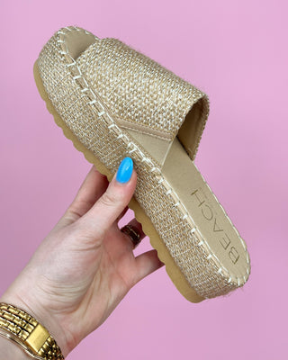 Gold glittery slipper held by a hand with blue nail polish against a pink background