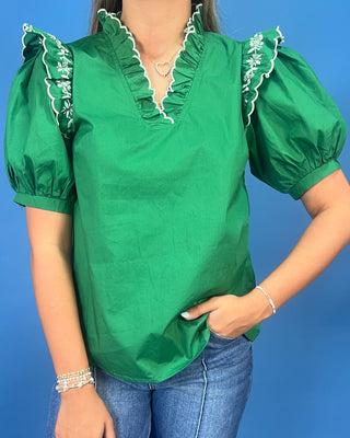 A woman wearing a bright green short-sleeve blouse with ruffled trim and puff sleeves, paired with blue jeans, against a solid blue background.