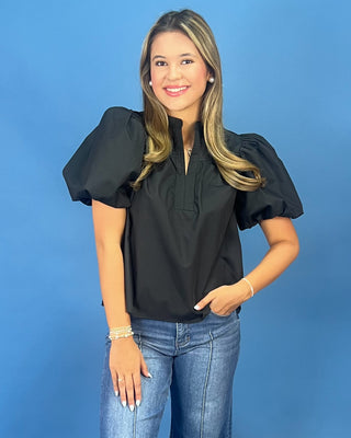 A woman modeling a black puff-sleeve blouse with a split neckline, paired with blue jeans, against a solid blue studio background.