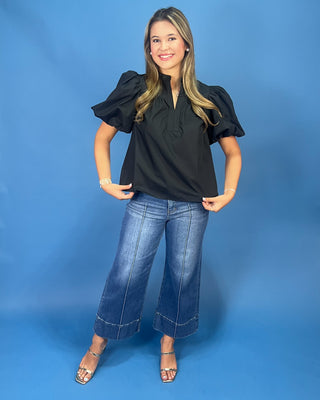 A woman modeling a black puff-sleeve blouse with a split neckline, paired with wide-leg blue jeans and metallic heeled sandals, standing against a solid blue studio backdrop.
