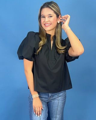 A woman modeling a black puff-sleeve blouse with a split neckline, paired with blue jeans, set against a solid blue studio background.