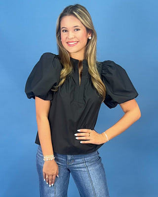 A woman modeling a black puff-sleeve blouse with a split neckline and front slit, paired with blue jeans, against a solid blue studio background.