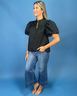 A woman modeling a black puff-sleeve blouse with a split neckline, paired with wide-leg blue jeans and metallic sandals, standing against a solid blue studio backdrop.