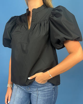 A woman wearing a black puff-sleeve blouse with a split neckline and gathered chest detail, paired with blue jeans, against a solid blue background.