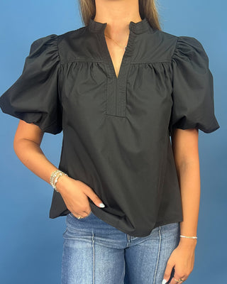 A woman wearing a black short-sleeve blouse with puff sleeves and a gathered mock neck, paired with blue jeans, against a solid blue background.