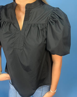 A woman wearing a black short-sleeve blouse with puff sleeves, a split neckline, and gathered detailing, paired with blue jeans against a solid blue background.