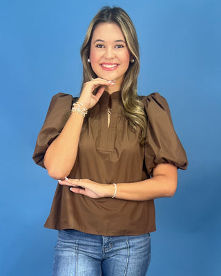 A woman modeling a brown short-sleeve blouse with puffed sleeves and a split neckline, paired with blue jeans, against a solid blue background.