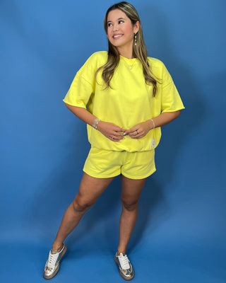 A woman modeling a Lulu Mac bright yellow oversized short-sleeve t-shirt and matching yellow shorts against a solid blue studio backdrop.