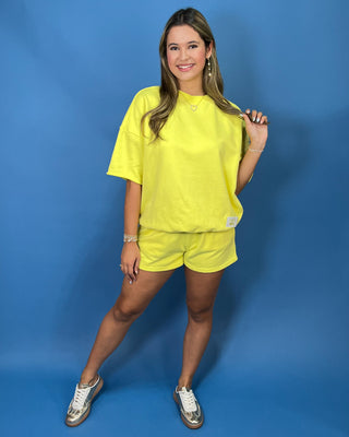 A woman modeling a Lulu Mac bright yellow oversized short-sleeve t-shirt and matching yellow shorts against a solid blue studio backdrop.