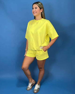 A woman modeling a Lulu Mac bright yellow oversized short-sleeve t-shirt and matching yellow shorts against a solid blue studio backdrop.