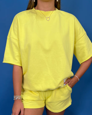 A woman modeling a Lulu Mac bright yellow oversized short-sleeve t-shirt and matching yellow shorts against a solid blue studio backdrop.
