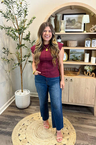 Woman in a burgundy top and blue jeans standing in a room with a plant and wooden cabinet.