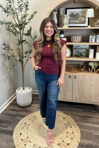 Woman in a maroon top and blue jeans standing in a room with a plant and shelves.