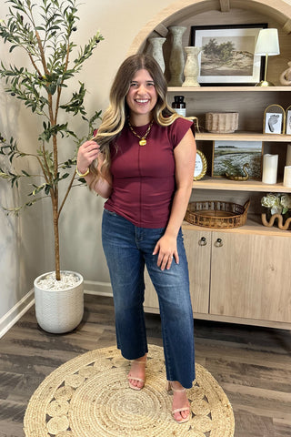 Woman in a maroon top and blue jeans standing in a room with a plant and wooden cabinet.