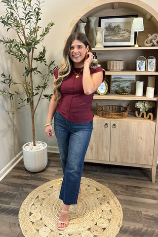 Woman in a maroon top and blue jeans standing in a room with a plant and wooden cabinet.