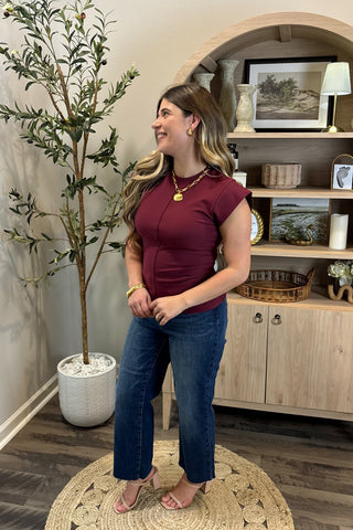 Woman in a burgundy top and blue jeans standing in a room with a plant and wooden cabinet.