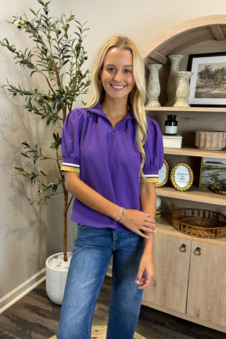 Woman wearing a purple polo shirt with yellow accents standing in a room with a plant and wooden shelf.