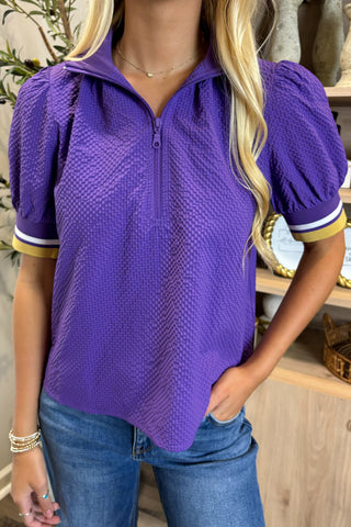 Woman wearing a purple textured shirt with short sleeves and blue jeans indoors.