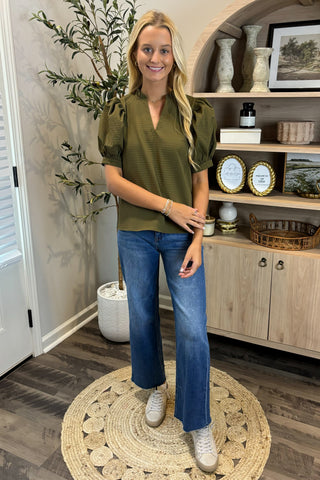Woman in a green top and blue jeans standing in a room with a plant and shelves.