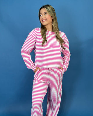 A woman modeling a lulu mac pink and white striped two-piece lounge set consisting of a long-sleeve top and matching pants, standing against a solid blue studio backdrop.