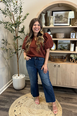 Woman in a rust-colored top and blue jeans standing in a room with a plant and wooden cabinet.
