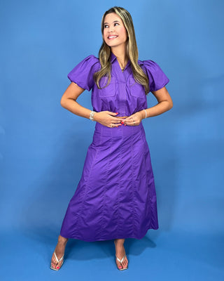 A woman modeling a purple midi dress with short puff sleeves, a gathered waist, and a crinkled texture, standing against a s
