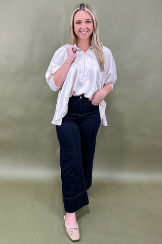 Woman wearing a white blouse and dark jeans against a plain background