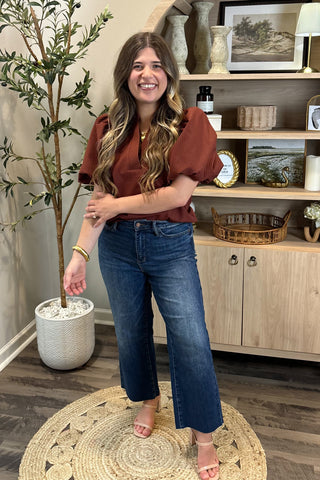 Woman in a rust-colored top and blue jeans standing in a room with a plant and wooden shelves.