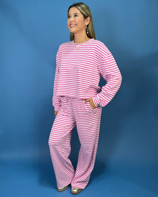 A woman modeling a lulu mac pink and white striped two-piece lounge set consisting of a long-sleeve top and matching pants, standing against a solid blue studio backdrop.