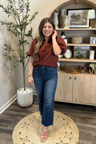 Woman in a brown top and blue jeans standing in a room with a plant and wooden cabinet.