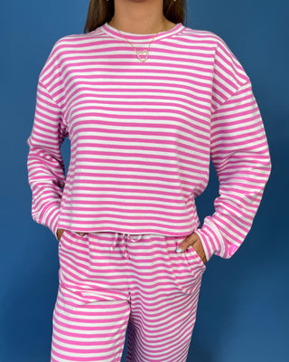 A woman modeling a lulu mac pink and white striped two-piece lounge set consisting of a long-sleeve top and matching pants, standing against a solid blue studio backdrop.