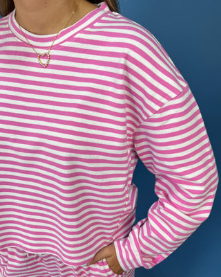 A woman modeling a lulu mac pink and white striped two-piece lounge set consisting of a long-sleeve top and matching pants, standing against a solid blue studio backdrop.