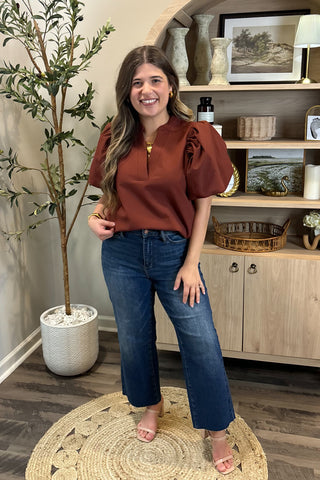 Woman in a rust-colored blouse and blue jeans standing in a room with a plant and wooden cabinet.