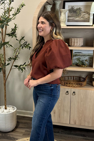 Woman in a rust-colored blouse and blue jeans standing in a room with a plant and wooden cabinet.