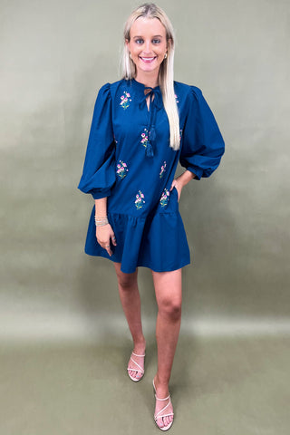 Woman wearing a blue dress with floral embroidery against a plain background
