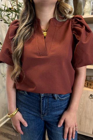 Person wearing a rust-colored top with ruffled sleeves and blue jeans indoors.